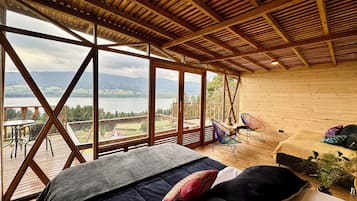 A spacious room with a large bed, two chairs, and a view of a lake and mountains through large windows.