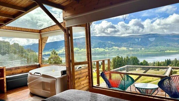 A wooden cabin with a large window offering a view of a lake and mountains. Inside, there is a bed, a small table, and a chair. - Neusa Hills Eco Hotel Boutique (Tausa)