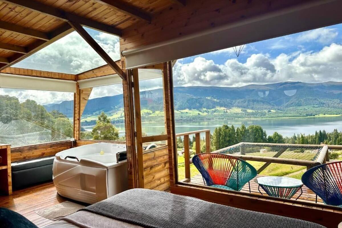 Eine Holzhütte mit einem großen Fenster, das einen Blick auf einen See und Berge bietet. Innen befinden sich ein Bett, ein kleiner Tisch und ein Stuhl.