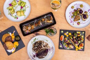 Verschiedene Gerichte auf einem Holztisch, darunter Tacos, eine schwarze, rechteckige Schale und ein Teller mit einem gegrillten Gericht.