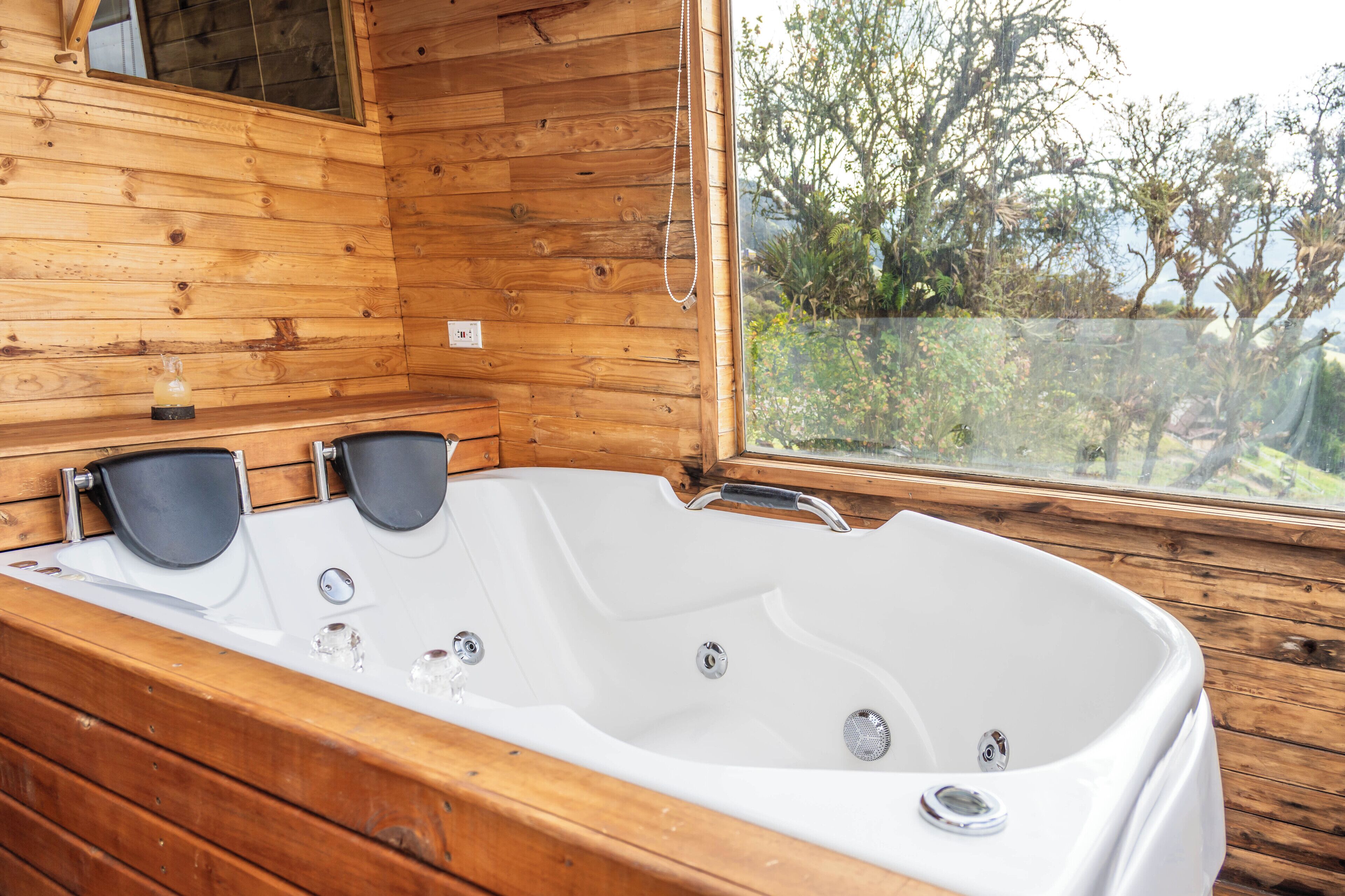A wooden cabin with a large white bathtub, a window with a view of trees, and a small shelf with a bottle.