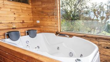 A wooden cabin with a large white bathtub, a window with a view of trees, and a small shelf with a bottle.
