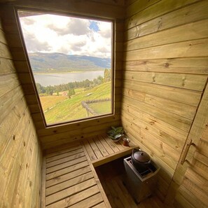 Eine Hütte mit großem Fenster mit Blick auf einen See und Berge. Die Hütte hat einen Holzboden und einen kleinen Tisch mit einem Topf und einer Tasse.