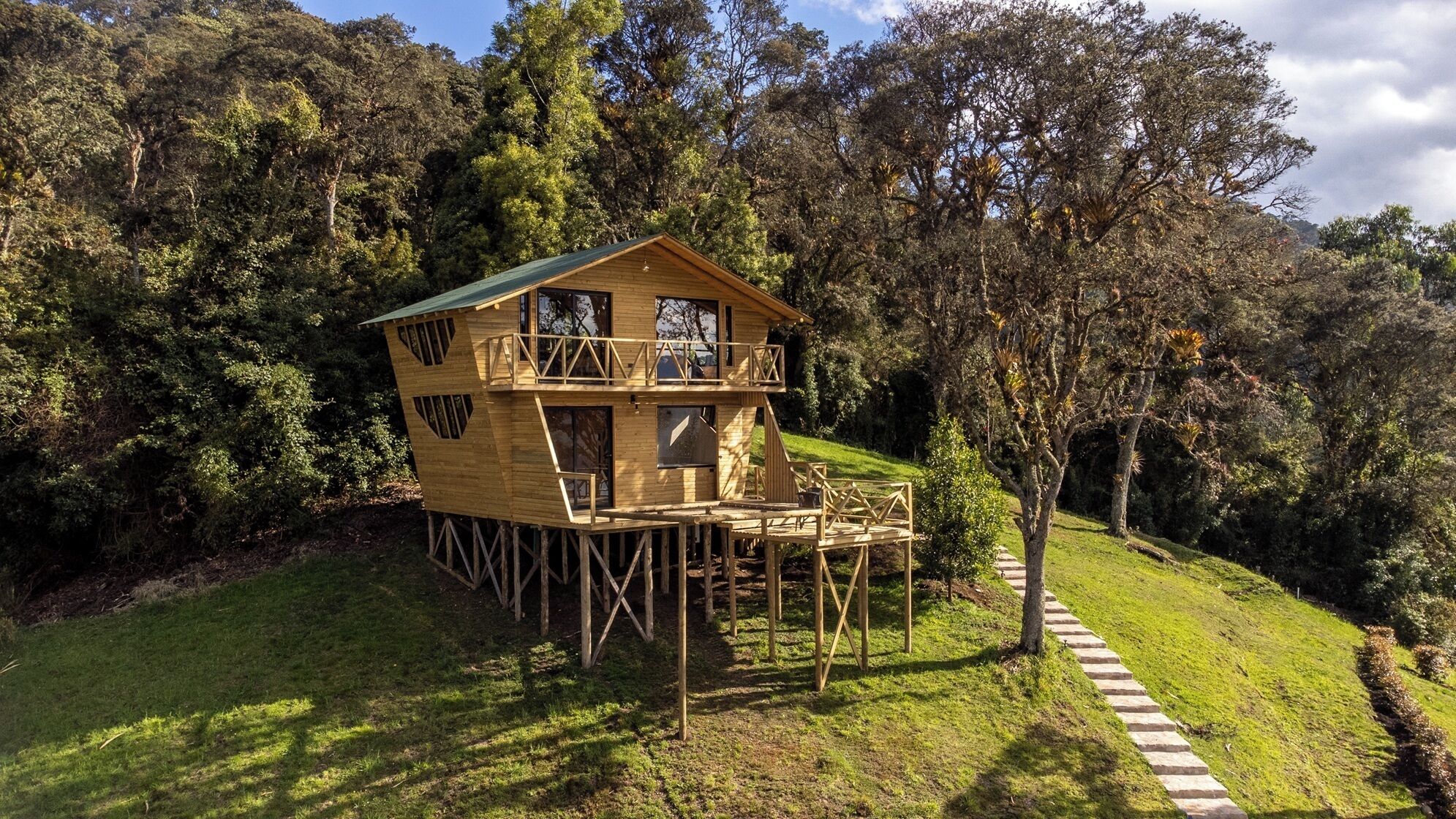 Una cabaña de madera de dos pisos con balcón, rodeada de abundante vegetación y un camino de piedra.