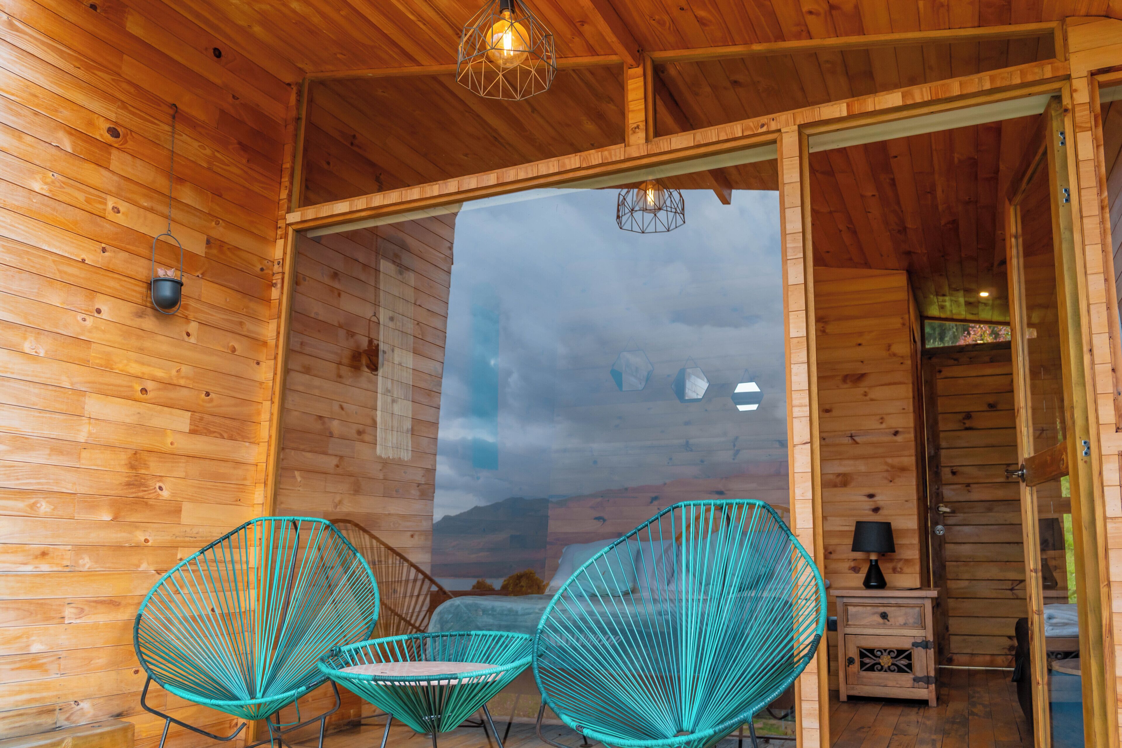 Interior de uma cabana de madeira com uma grande porta de vidro que dá para o exterior, duas cadeiras turquesa e uma pequena mesa de cabeceira com uma lâmpada.
