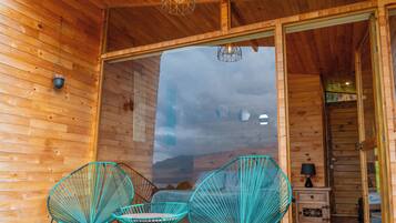 Interior de uma cabana de madeira com uma grande porta de vidro que dá para o exterior, duas cadeiras turquesa e uma pequena mesa de cabeceira com uma lâmpada.