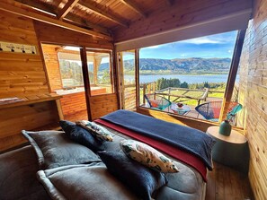 Une chambre dans un chalet en bois, avec un lit, une fenêtre donnant sur un lac et des montagnes, et un balcon avec des chaises.