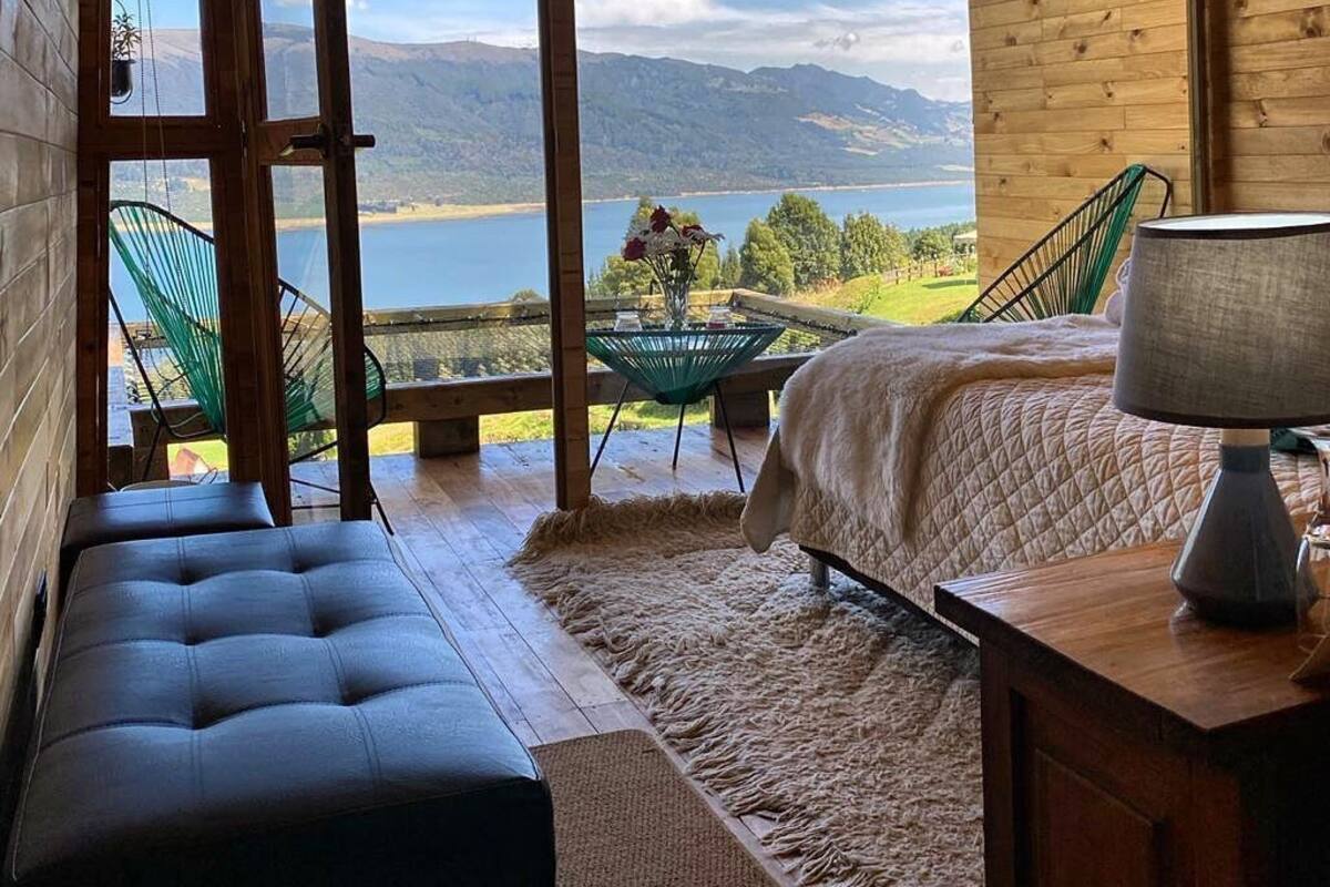 Ein Zimmer in einer Holzhütte mit einem Bett, einer Bank, einem Tisch und Blick auf einen See und Berge.