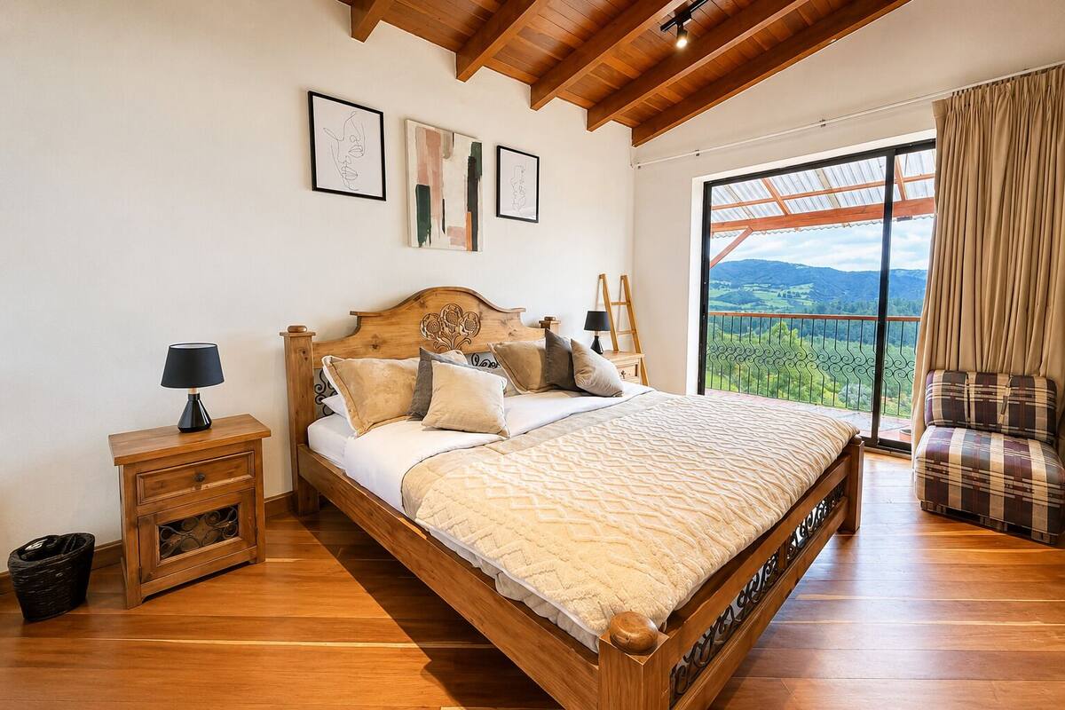 Ein Schlafzimmer mit einem hölzernen Bett, einem Nachttisch, einem Sessel und einem großen Fenster mit Aussicht.