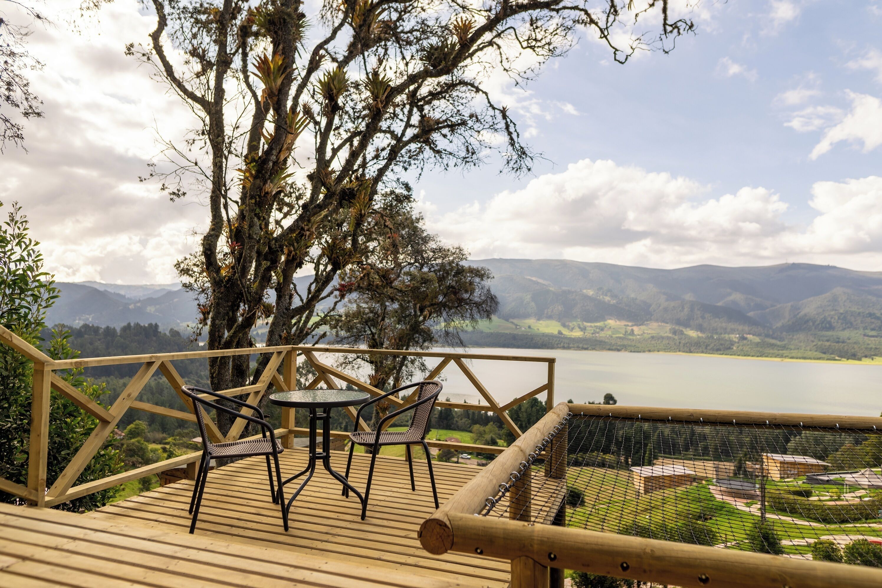 Um deck de madeira com duas cadeiras e uma mesa, com vista para um lago e montanhas.
