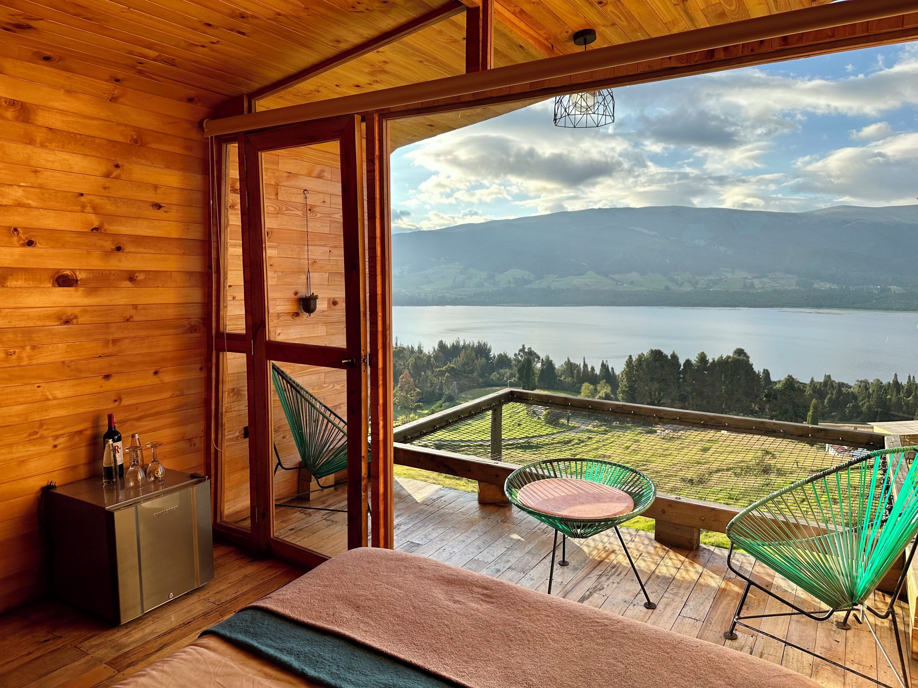 A wooden cabin room with a large window overlooking a lake and mountains.