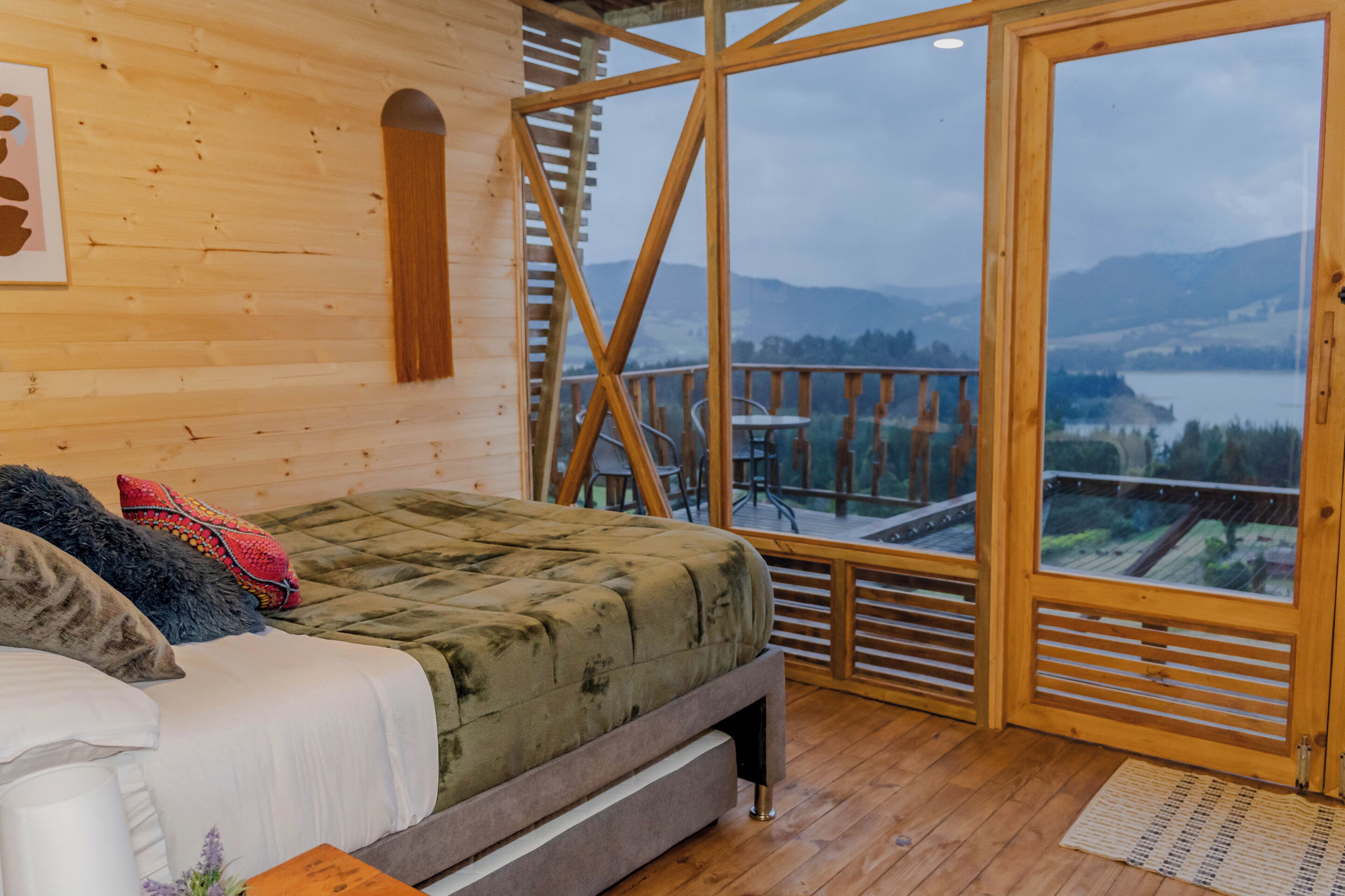 A wooden cabin room with a bed, a view of a lake, and a balcony with a railing.