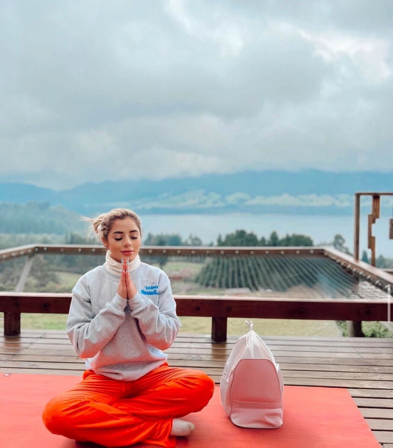 A person sitting on a red mat in a meditative pose on a wooden deck with a scenic view of mountains and a body of water in the background.