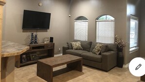 Ein Wohnzimmer mit einem Flachbildfernseher, einem Holztisch, einem grauen Sofa mit gemusterten Kissen und einem Deckenventilator.