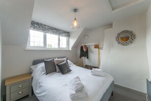 Ein ordentlich bezogenes Bett mit Kissen und einem gefalteten Handtuch, ein Nachttisch, ein Fenster mit Vorhang und ein an der Wand befestigter Spiegel.