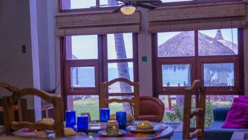 A dining area with a table set for two, large windows with a view of a thatched-roof structure, and a ceiling fan.
