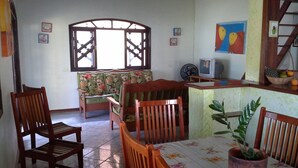 A dining area with wooden chairs and a table, a window with a wooden frame, a television, and a fan on a counter. - House for Season in closed condominium (Caraguatatuba)