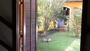 An open door leading to a patio with a tiled floor and a grassy area with trees and a yellow wall. - House for Season in closed condominium (Caraguatatuba)