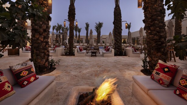 A lounge area with a central fire feature, surrounded by sofas and chairs, palm trees, and outdoor lighting. - Kedma by Isrotel Design (Ramat Negev)