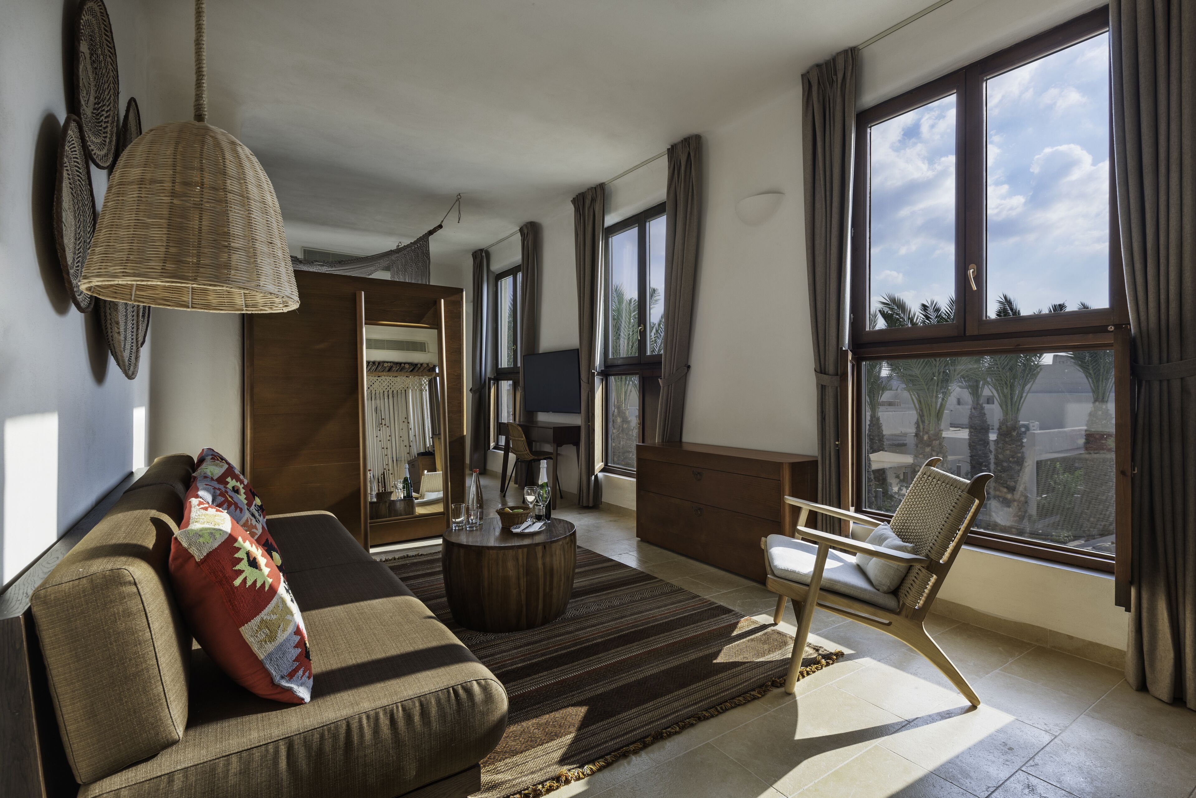 A modern living room with a sofa, armchair, and a large window offering a view of palm trees.
