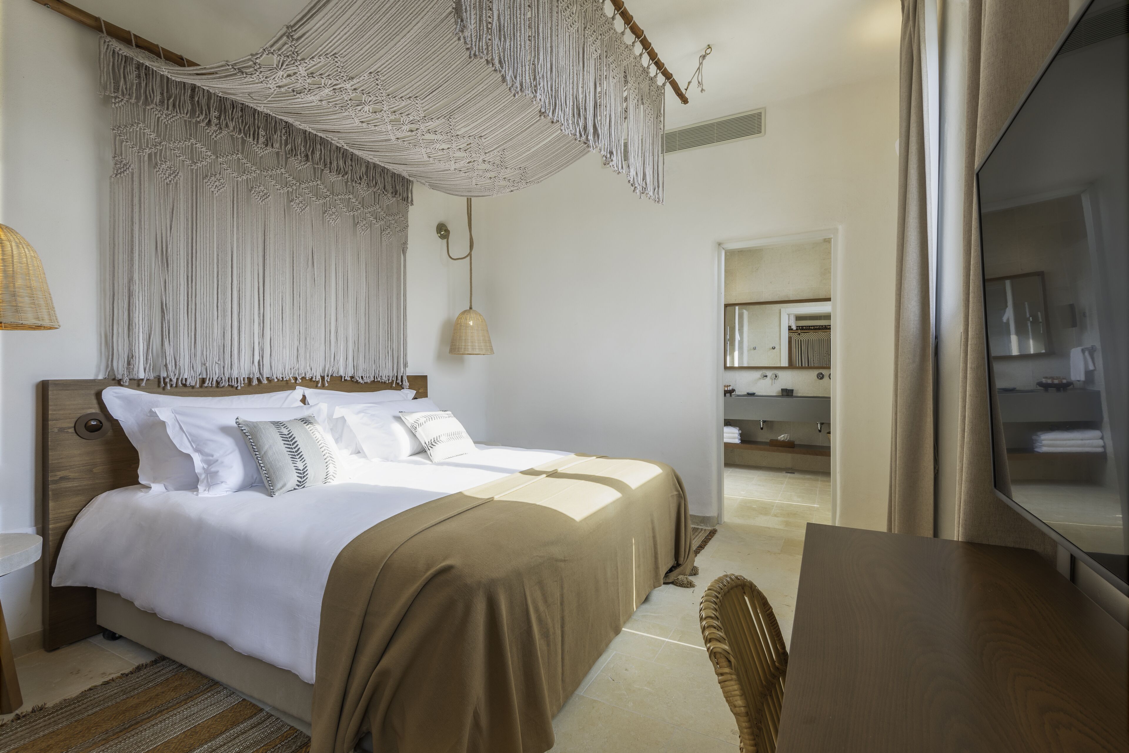 A modern hotel room with a large bed, a hanging macrame piece, a wooden nightstand, and a view of the bathroom.