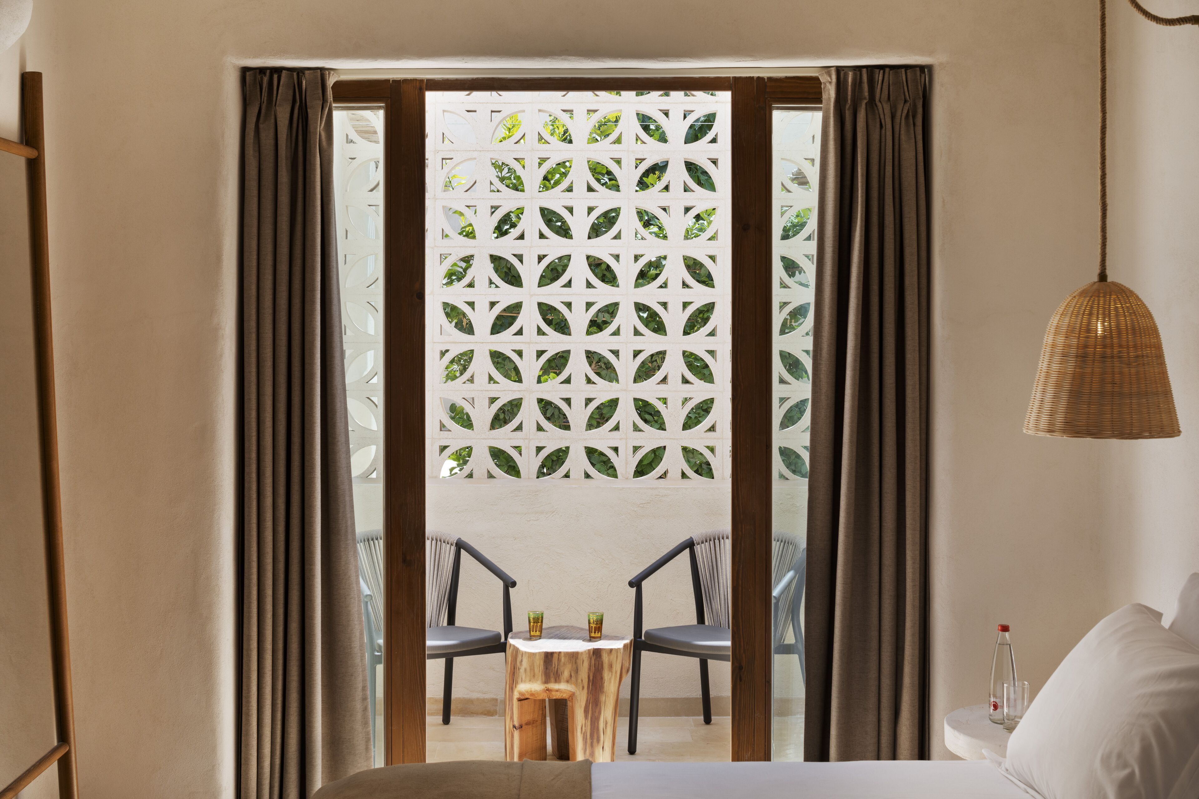 Habitación de hotel con cama, mesita de noche con una botella y vistas a través de una ventana con cortinas.