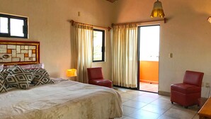 Una camera da letto con un letto, due poltrone rosse, un tavolo in legno, una lampada a parete e vista tramite una porta scorrevole.