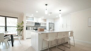A modern kitchen with white cabinetry, a central island with bar stools, a dining area with a table and chairs, and a large window with blinds. - Luxury Villa Miami Shores (Miami Shores)
