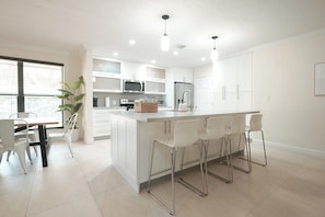 A modern kitchen with white cabinetry, a central island with bar stools, a dining area with a table and chairs, and a large window with blinds.