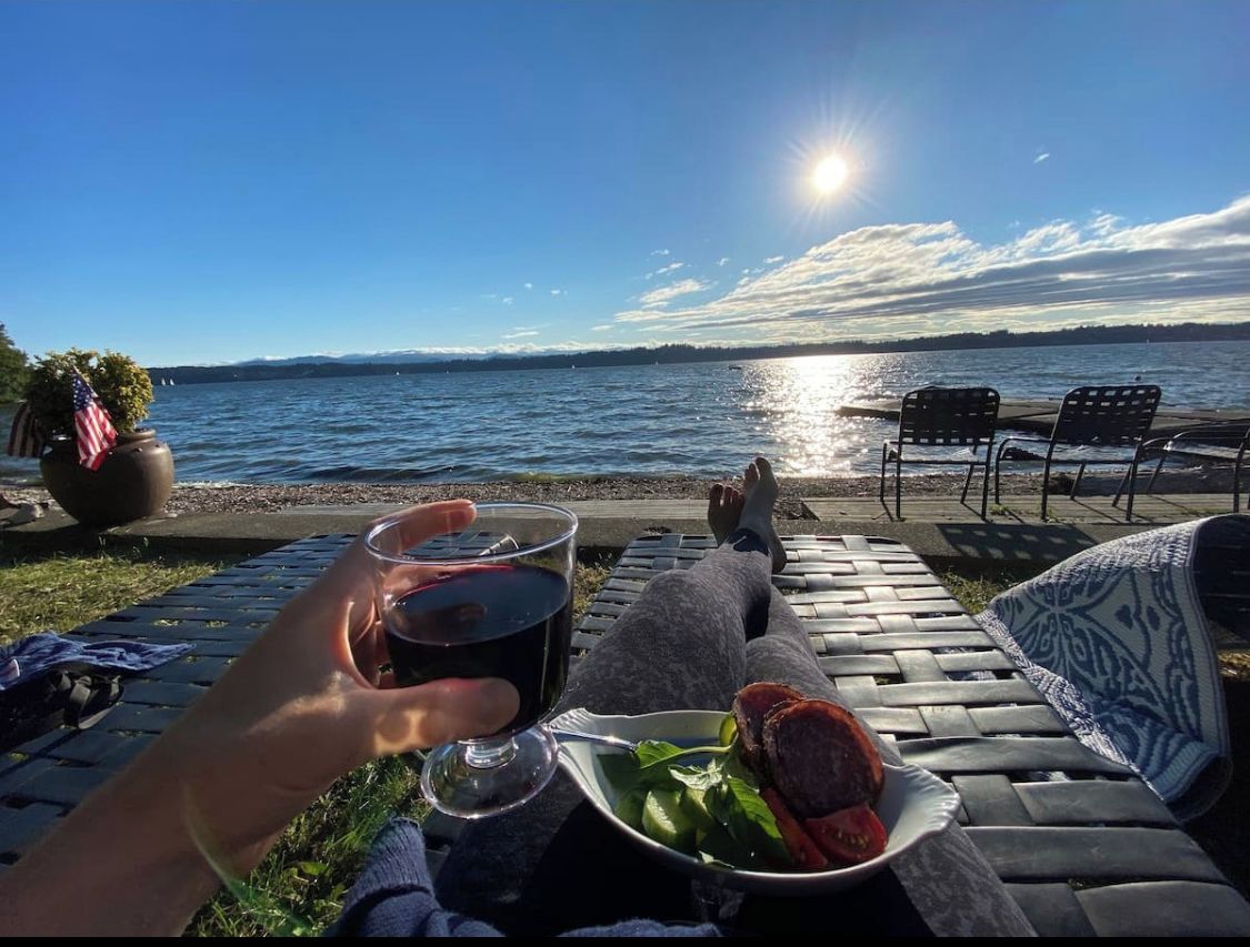 A waterfront patio with a view of the ocean, a table with a plate of food, and a glass of drink.