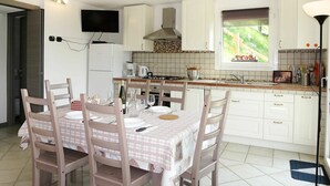 Une cuisine avec des meubles blancs, une table à manger recouverte d’une nappe à carreaux et une fenêtre donnant sur un paysage verdoyant.