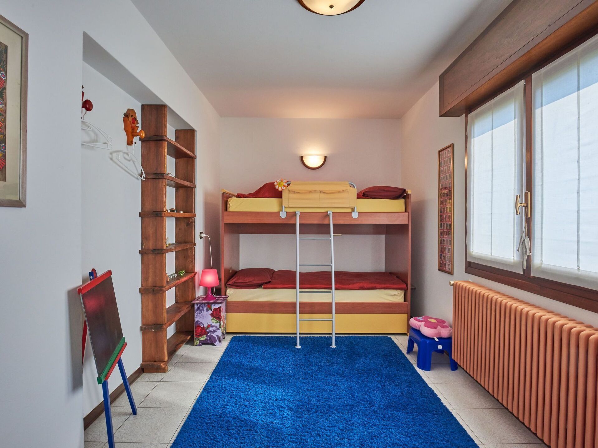Camera con letto a castello, tappeto blu, scala in legno, termosifone e finestra con persiane.