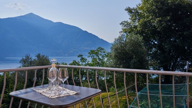 Une table avec trois verres à vin et une bouteille, placée sur un balcon donnant sur une montagne et un lac.