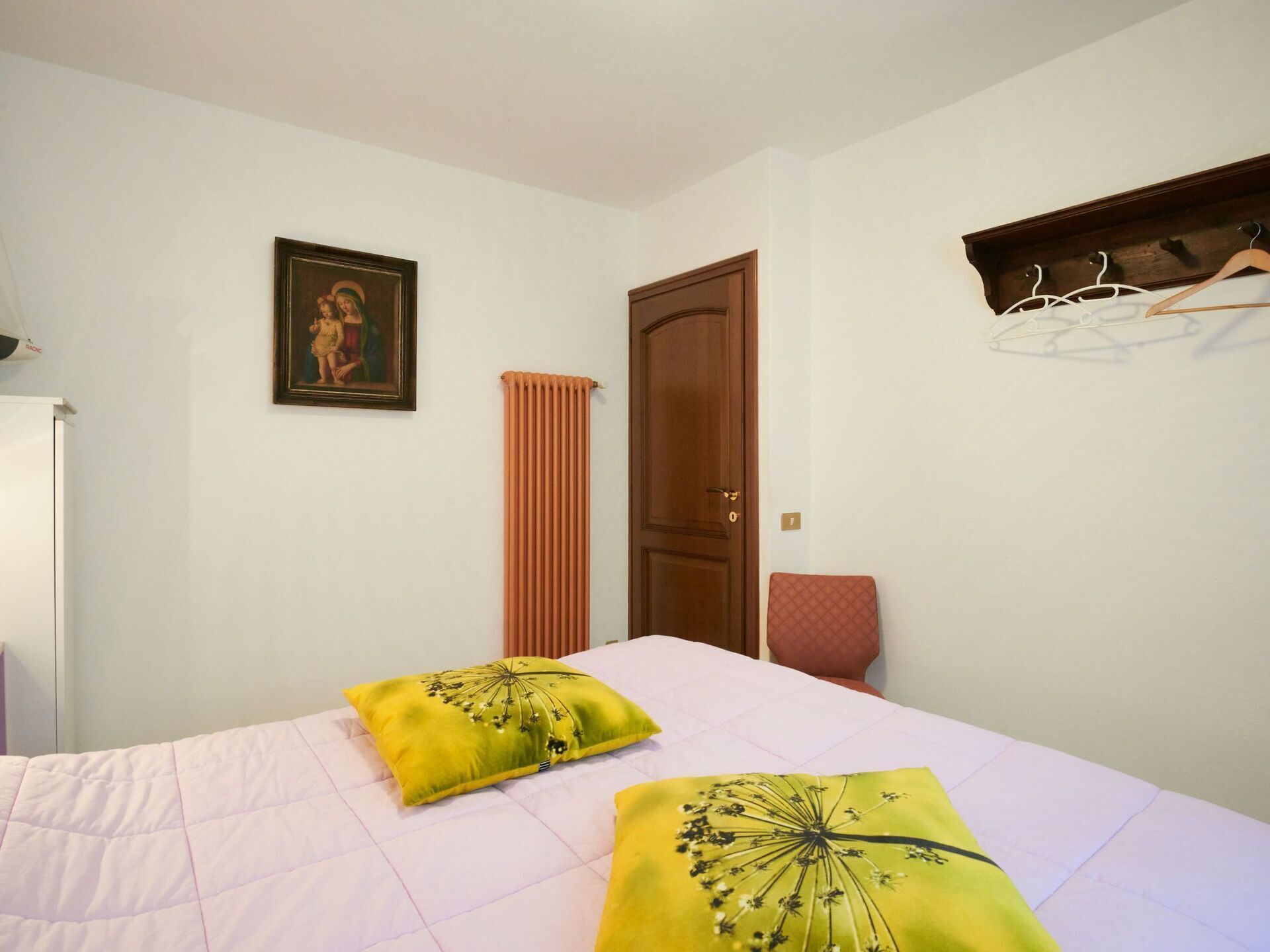 A bedroom with a bed, two pillows, a radiator, a door, a coat rack, and a framed picture on the wall.