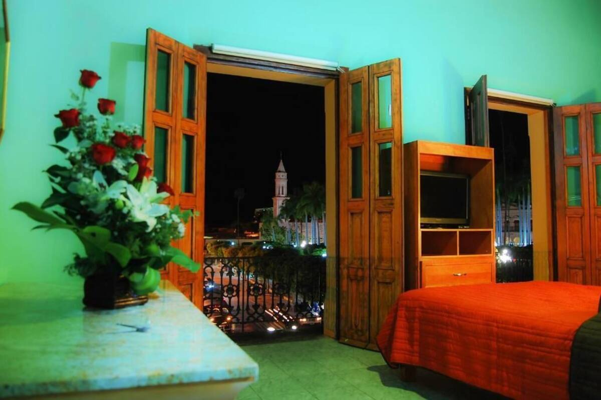A room with an open door revealing a night view of a cityscape, a bed with an orange bedspread, a television on a wooden stand, and a vase of flowers on a table.