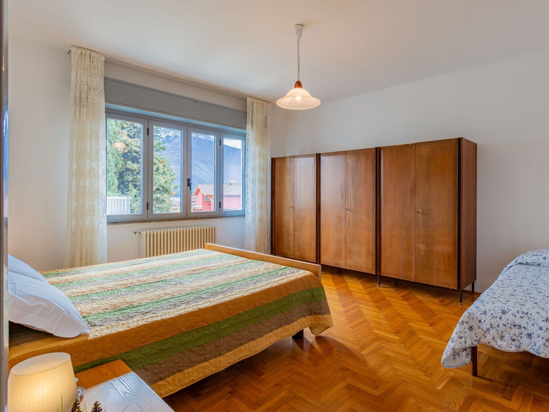 Une chambre à coucher avec un lit en bois, une fenêtre avec des rideaux, un radiateur, une armoire et un luminaire suspendu.