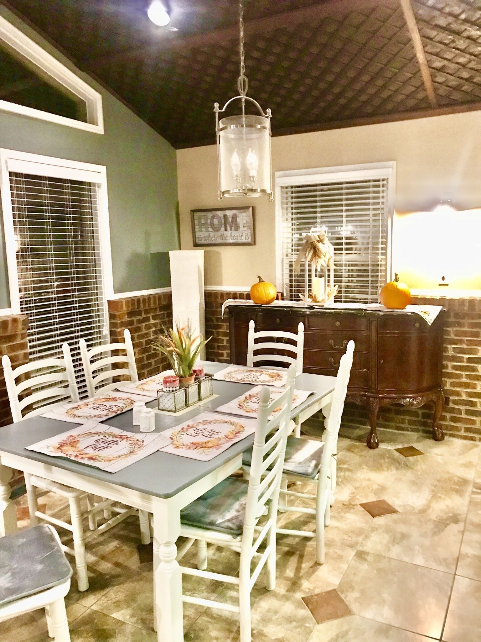 A dining area with a table set for four, white chairs, a chandelier, a brick wall, and a window with blinds.