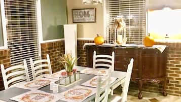 A dining area with a table set for four, white chairs, a chandelier, a brick wall, and a window with blinds.