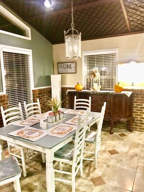 A dining area with a table set for four, white chairs, a chandelier, a brick wall, and a window with blinds.