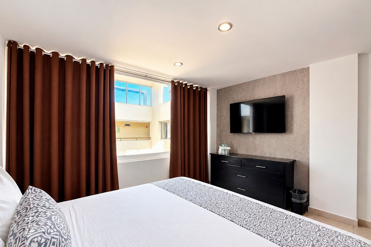 Een hotelkamer met een bed, een aan de muur bevestigde televisie, een dressoir en een raam met gordijnen.