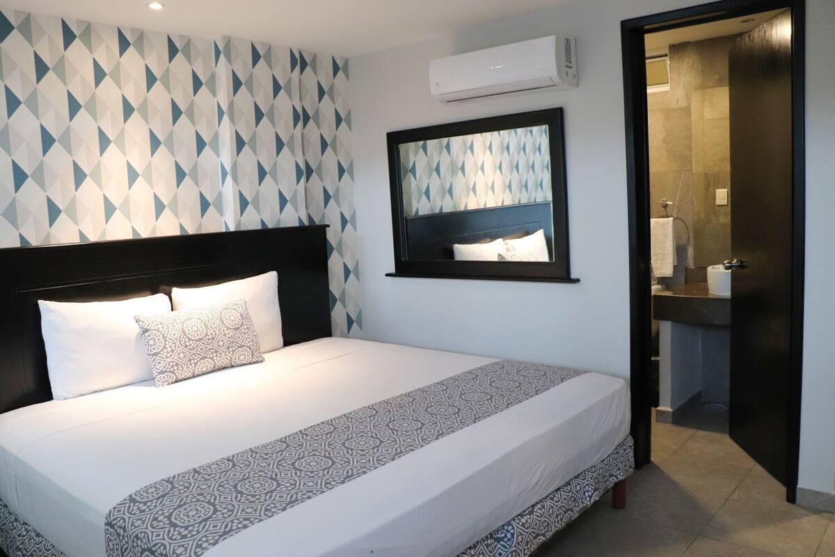A hotel room with a bed, a headboard, a wall-mounted air conditioner, a bathroom with a sink and mirror, and a patterned wall.