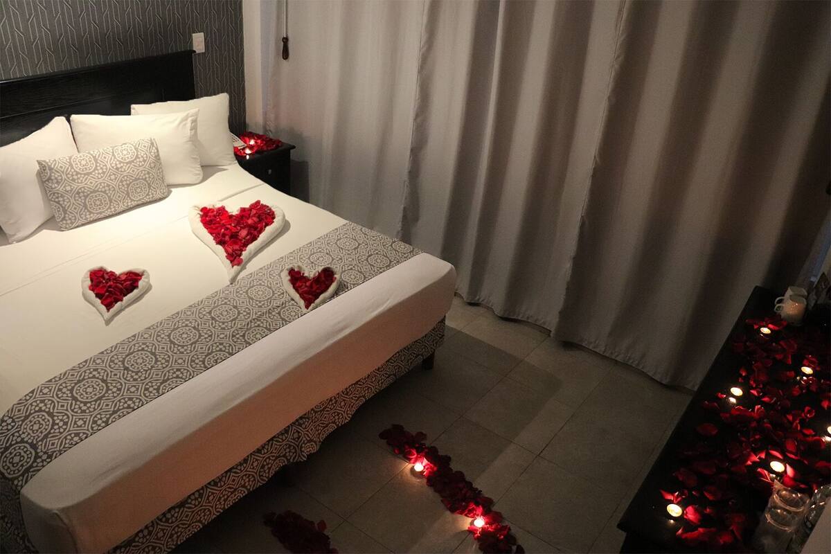 A neatly made bed with patterned bedding, a bedside table with a remote control, and a decorative heart-shaped arrangement of red roses on the bed.