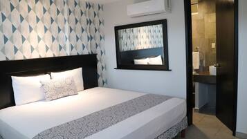 A hotel room with a bed, a headboard, a patterned wall, an air conditioning unit, and a bathroom visible through an open door.
