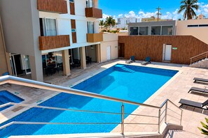 Una piscina con cubierta de madera, sillas de descanso y una escalera que lleva a un edificio con balcones.