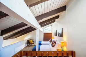 A bedroom with a sloped ceiling, a bed with a blue accent, a bedside lamp, and a window with blinds.