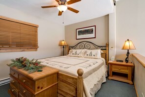 Un dormitorio con una cama de madera, dos mesitas de noche con lámparas, un ventilador de techo, una ventana con persianas y un cuadro en la pared.
