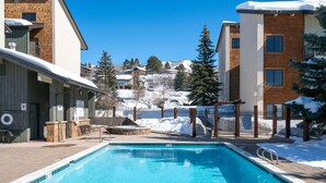 A swimming pool with a clear blue water, surrounded by buildings with snow on the roof and trees. - Charming Studio + Loft Condo | Walk to Gondola | Pool, Hot Tubs and Views! (Steamboat Springs)