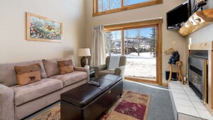 A living room with a sofa, a coffee table, a fireplace, a television, and a large window with a view of a snowy landscape. - Charming Studio + Loft Condo | Walk to Gondola | Pool, Hot Tubs and Views! (Steamboat Springs)