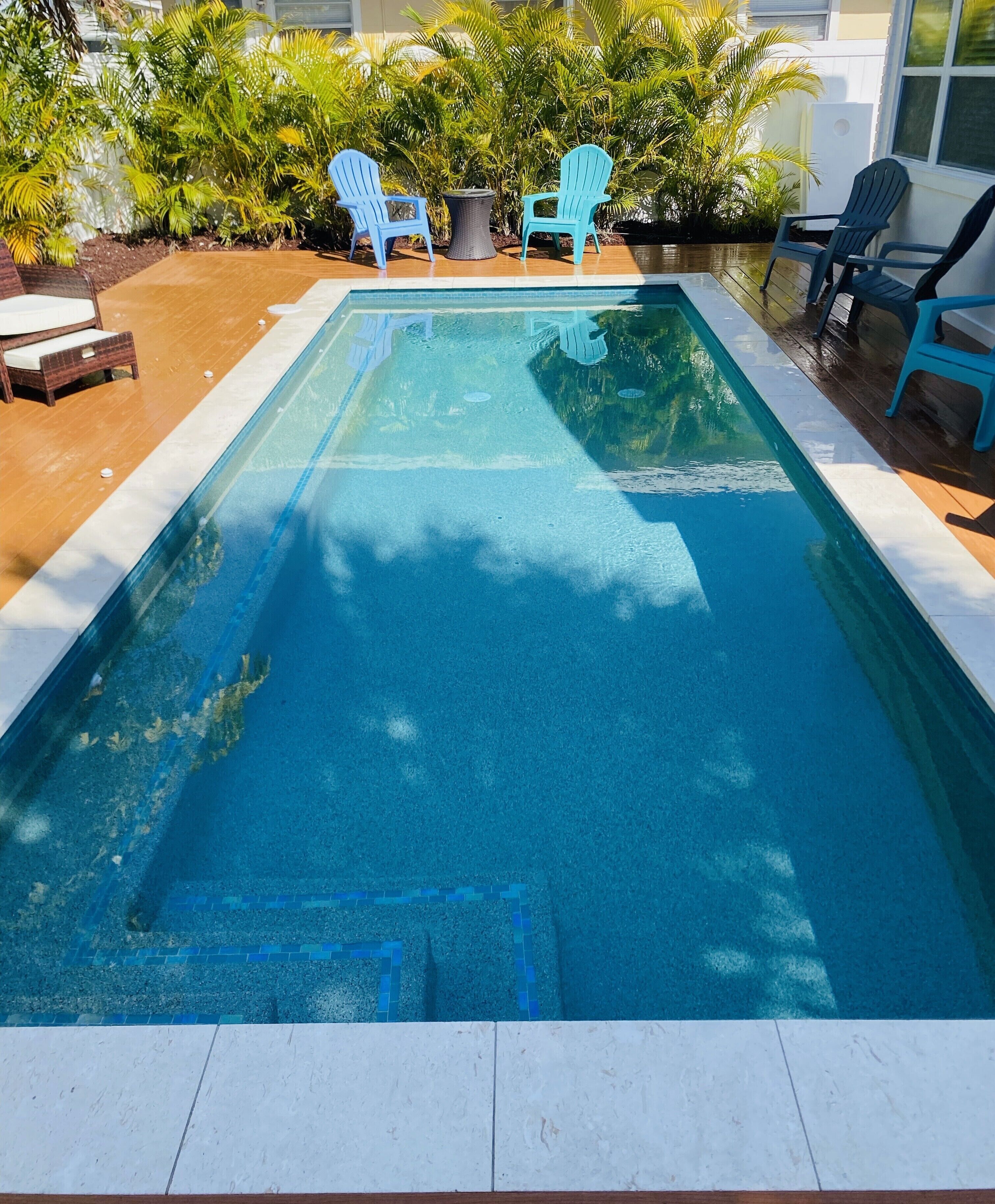 Uma piscina retangular com borda ladrilhada, rodeada por um deck de madeira. Há várias cadeiras Adirondack azuis e uma pequena mesa redonda com uma planta.