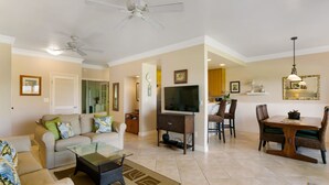 A spacious living area with a sofa, coffee table, and a dining table with chairs. A television is mounted on the wall, and there is a ceiling fan. - Deluxe Ocean View Ground Floor.... Renovated ..Walk to the Hyatt and Beach...A/C (Koloa)
